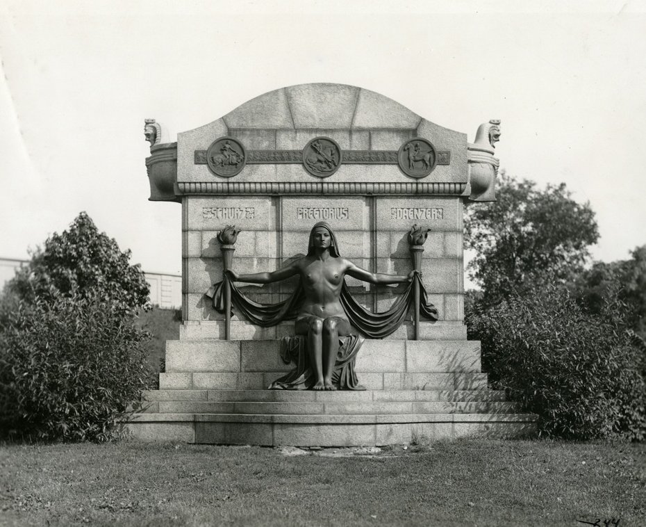 "THE NAKED TRUTH." (PREETORIUS-SCHURZ-DAENZER MONUMENT). Horizontal, black and white photograph showing a monument with a statue of a naked woman seated holding a touch in each outstretched hand. Three names above her head read: "Schurz. Preetorius. Daenzer." On either side of the monument are shrubs.